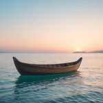 Um barco de madeira flutuando calmamente em um mar tranquilo ao amanhecer, com céu iluminado por tons dourados e rosados e montanhas ao fundo.