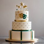 Bolo elegante decorado com detalhes verdes e dourados, com enfeites pequenos no topo, colocado sobre uma superfície refletiva com fundo neutro.